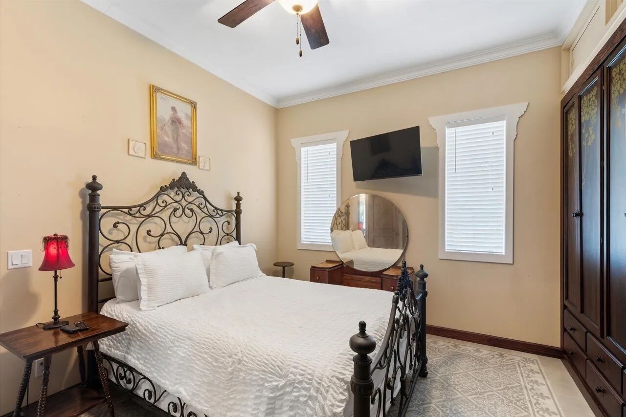 A bedroom with a bed, a bedside table with a lamp, a ceiling fan, a wall-mounted TV, and a window with blinds.