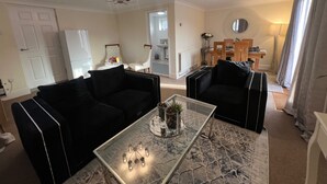 Una sala de estar con dos sofás negros, una mesa de centro de vidrio con objetos decorativos, una araña y un comedor al fondo.