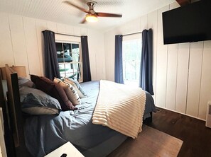 A bedroom with a bed, a ceiling fan, a window with curtains, and a flat-screen TV mounted on the wall.
