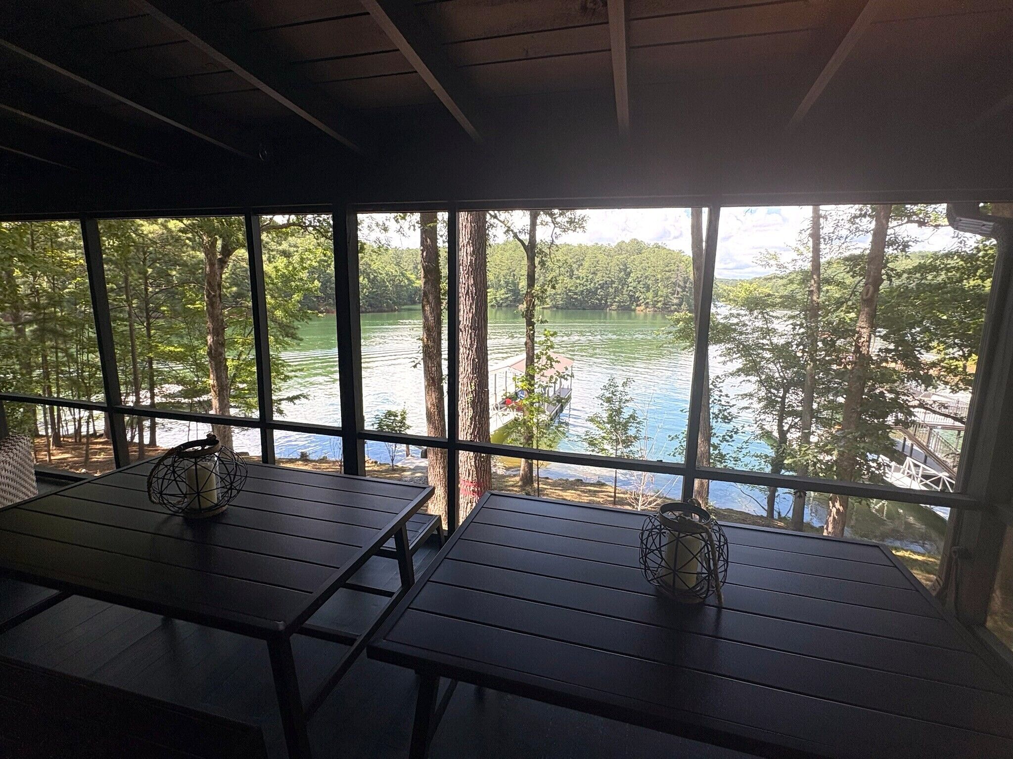 A covered outdoor seating area with tables and chairs, overlooking a lake surrounded by trees.