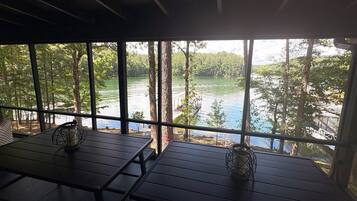 A covered outdoor seating area with tables and chairs, overlooking a lake surrounded by trees.