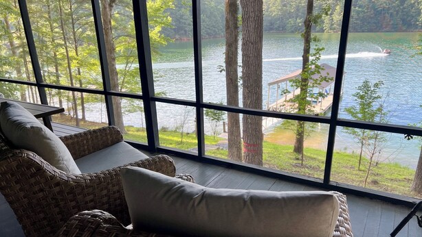 A wicker sofa with cushions, a wooden table, and a large window overlooking a lake and forest.