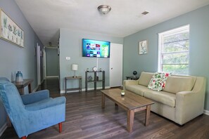 A living room with a blue armchair, a beige sofa with a floral throw, a wooden coffee table, and a flat-screen TV mounted on the wall.