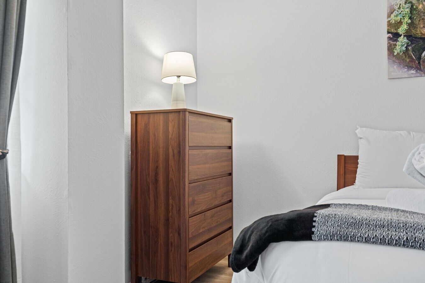 A bedroom with a wooden dresser, a bedside lamp, a bed with white bedding, and a painting on the wall.