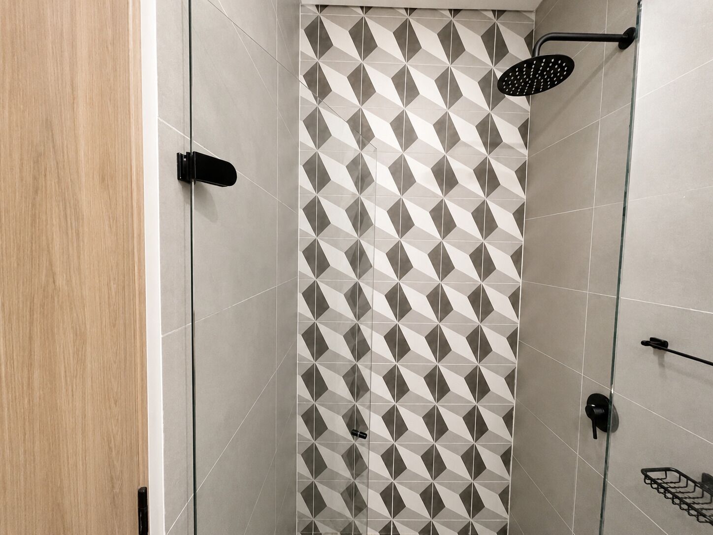 Un área de ducha con azulejos de diseño geométrico, un mamparo de vidrio y un cabezal de ducha negro.