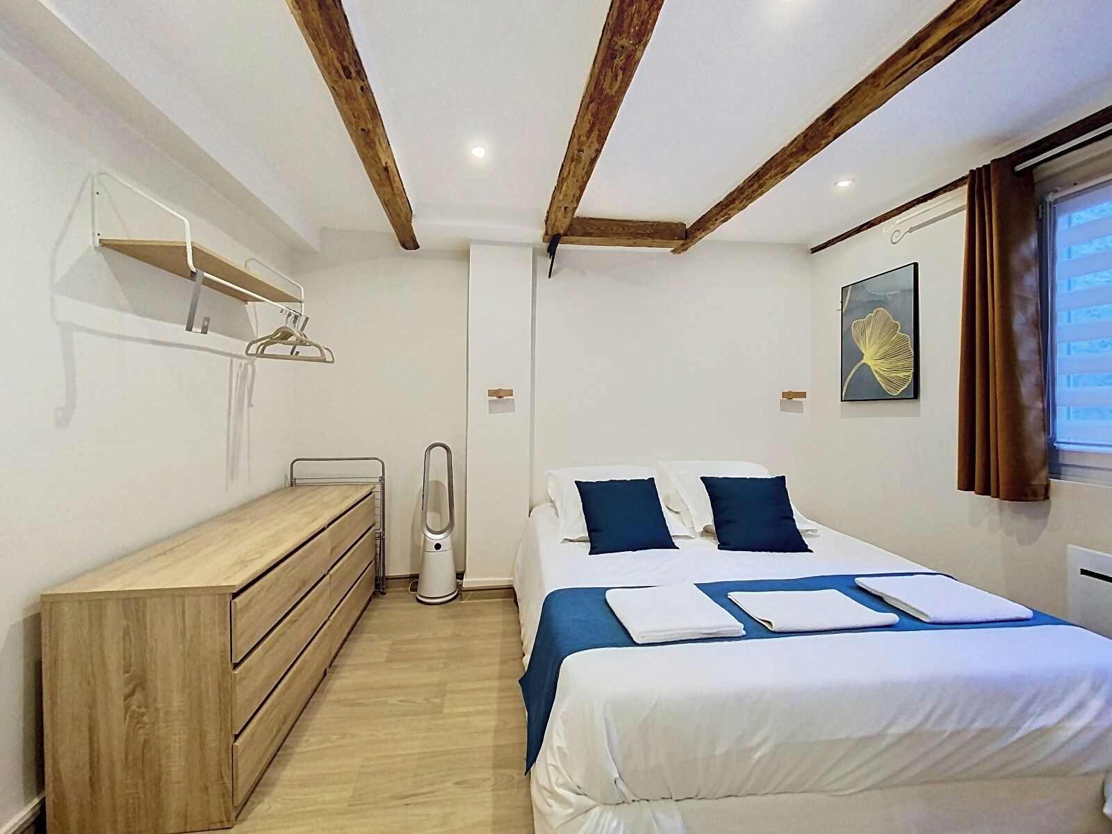Un dormitorio con cama, mesitas de noche, cómoda de madera, ventilador de techo y ventana con cortinas.
