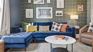 Un salon avec un canapé d’angle bleu, une table basse ronde et un mur orné d’un motif à carreaux, sur lequel sont accrochées des œuvres d’art encadrées.
