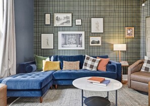 Un salon avec un canapé d’angle bleu, une table basse ronde et un mur orné d’un motif à carreaux, sur lequel sont accrochées des œuvres d’art encadrées.