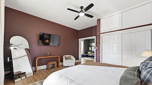 Une chambre à coucher comprenant un lit, un ventilateur de plafond, une télévision, un miroir, un bureau et une chaise.