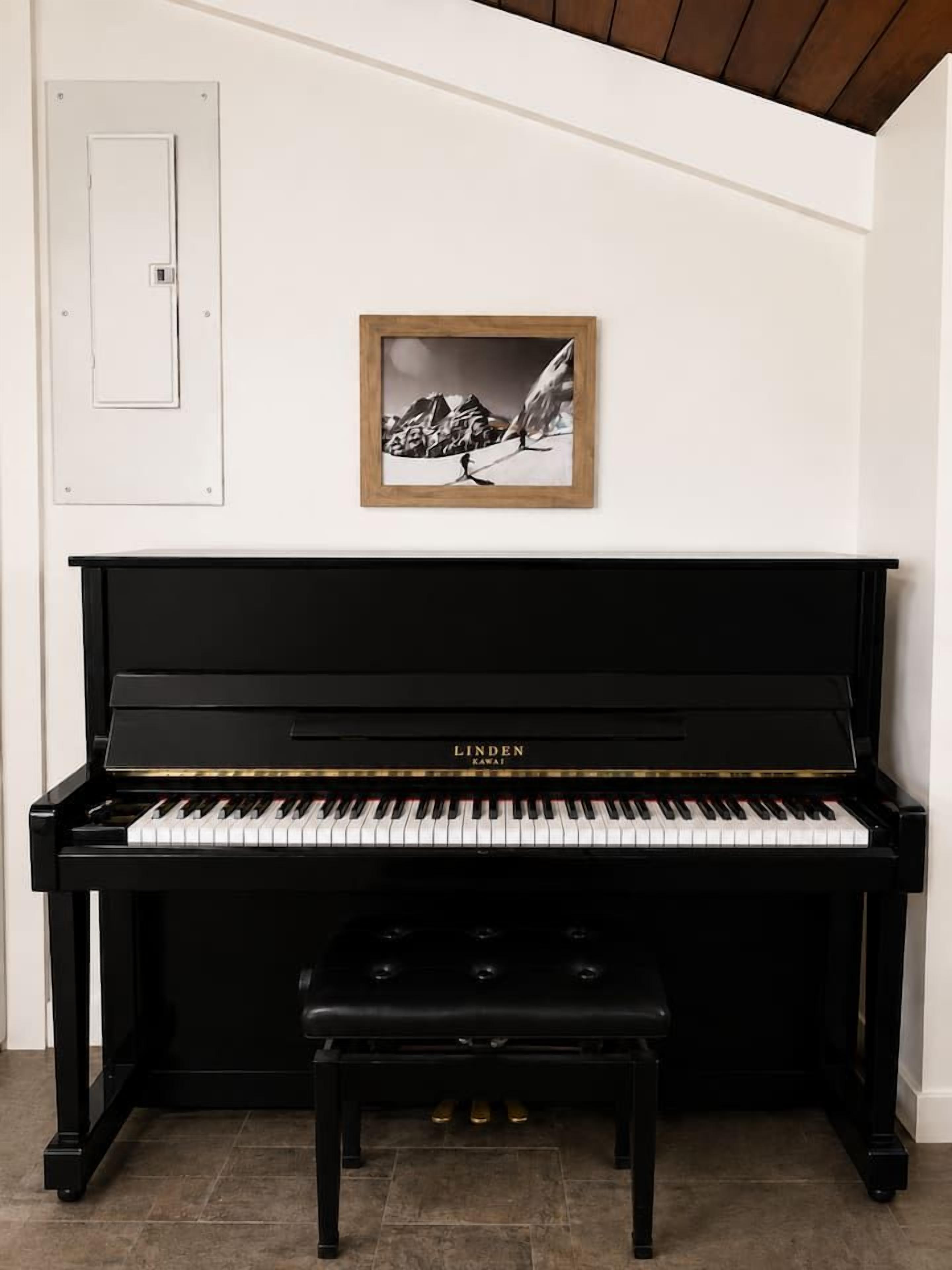 Un piano droit noir, avec un banc devant, un tableau encadré au mur et une porte blanche sur la gauche.