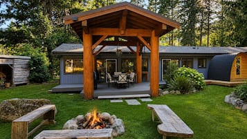 A wooden gazebo with a fire pit in the center, surrounded by a well-maintained lawn and picnic tables.