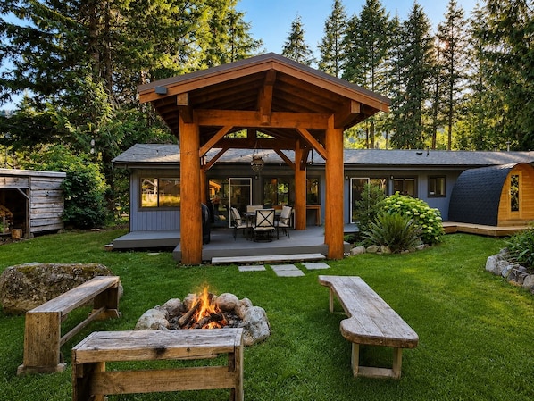A wooden gazebo with a fire pit in the center, surrounded by a well-maintained lawn and picnic tables.