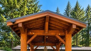 A wooden pergola structure with a patio area, outdoor seating, and a view of a house in the background. - Mountain Escape | Sauna & Outdoor fire Pit (Squamish)