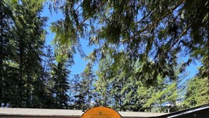 A wooden structure with a door, set in a grassy area with a building in the background. - Mountain Escape | Sauna & Outdoor fire Pit (Squamish)