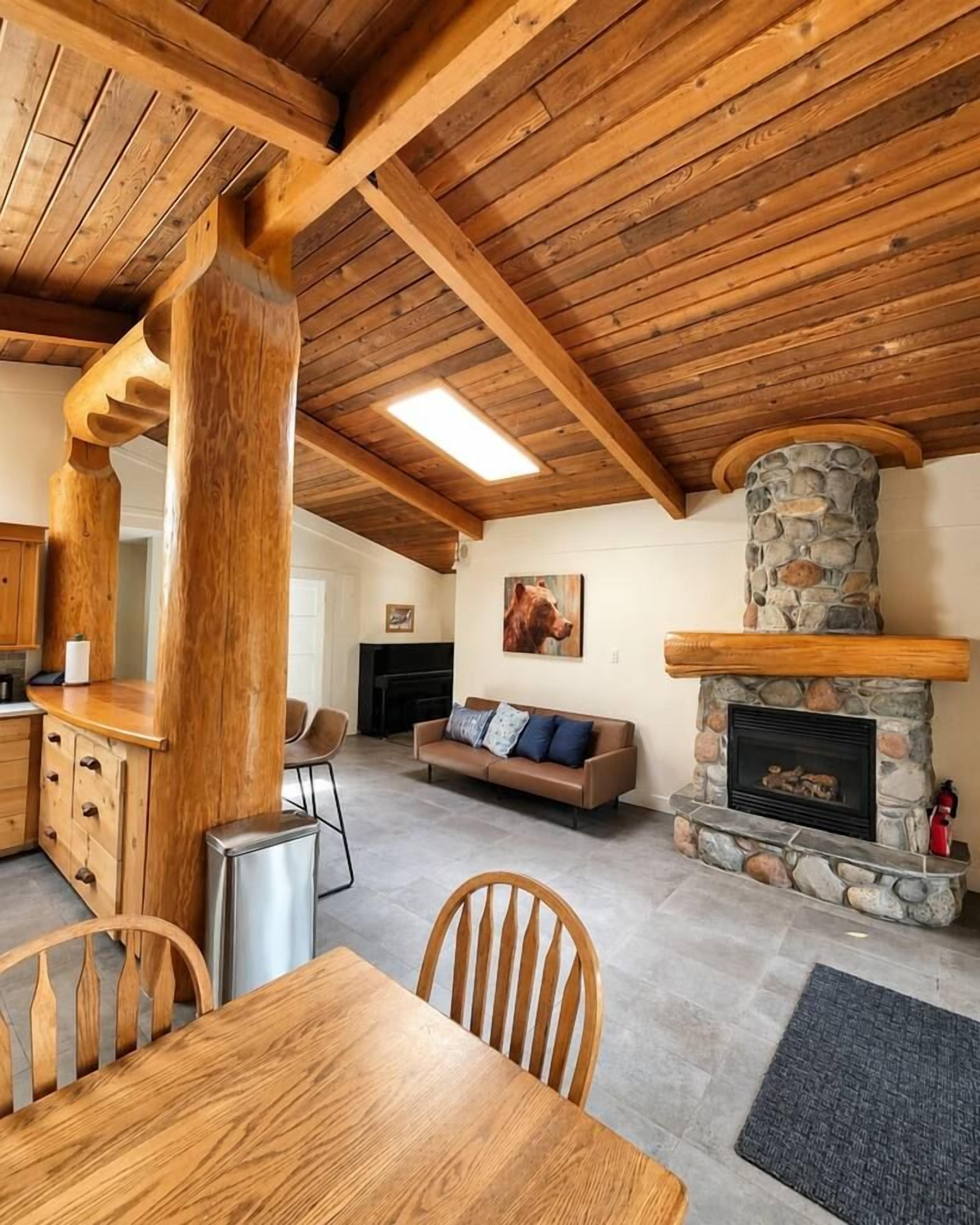 Un intérieur rustique avec une cheminée en pierre, des poutres apparentes en bois au plafond, un coin repas et un espace de vie comprenant un canapé et un tableau.