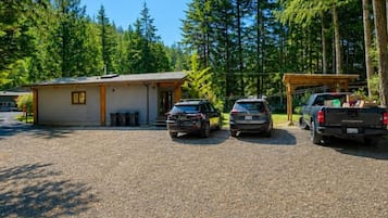 Un espace de stationnement recouvert de gravier, où sont garés trois véhicules, ainsi qu’un petit bâtiment et une forêt en arrière-plan.