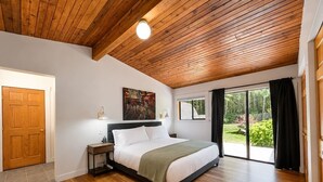 Ein Schlafzimmer mit Holzbalkendecke, einem Bett mit weißer Bettwäsche, einem Nachttisch, einem Gemälde an der Wand und Blick ins Freie durch eine Schiebetür.