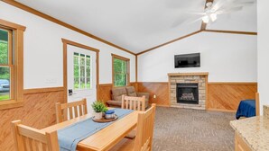 A room with a wooden dining table, chairs, a sofa, a fireplace, and a ceiling fan. - Whitetail Ridge Cabin 1 (Lexington)