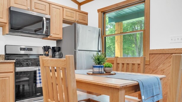 A kitchen with wooden cabinets, a stainless steel refrigerator, a microwave, and an oven. There is a window with a view of trees. - Whitetail Ridge Cabin 1 (Lexington)