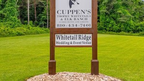 A wooden sign with a deer head logo, 'CUPPEN'S Trophy Whitetails & Elk Ranch' and contact number, 'Whitetail Ridge Wedding & Event Venue' on a grassy field. - Whitetail Ridge Cabin 1 (Lexington)