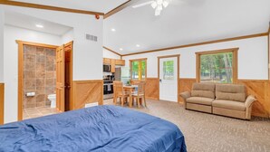 A room with a bed, a sofa, a dining table with chairs, a kitchen area, and a view of trees through the windows. - Whitetail Ridge Cabin 1 (Lexington)
