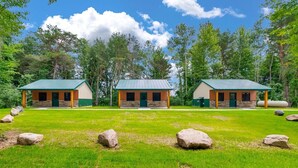 A lodge with three cabins, each with a green roof and stone walls, set in a grassy area with trees in the background. - Whitetail Ridge Cabins | Three Cabin Retreat (Lexington)