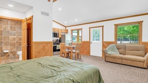 A room with a bed, a sofa, a dining table with chairs, a kitchen area, and a window with a view of trees.