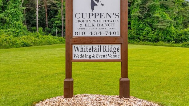 A wooden sign with a deer head logo, 'CUPPEN'S Trophy Whitetails & Elk Ranch' and contact number, 'Whitetail Ridge Wedding & Event Venue' on a grassy field.