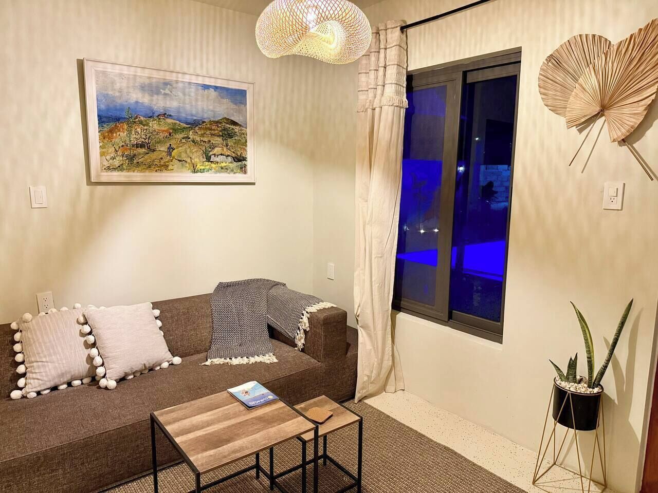 Uma sala de estar aconchegante com um sofá, uma mesa pequena, um quadro, uma janela com cortinas e um vaso de planta.
