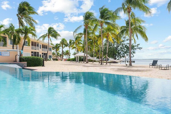 A pool with a beach area, palm trees, and a building with balconies.
