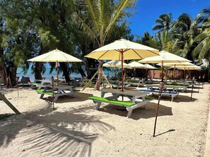 A beachside area with picnic tables and umbrellas, surrounded by palm trees and a view of the ocean. - Kai-Tastic 15B - 2BR Kaibo Condo with Pool and Beach (Rum Point)