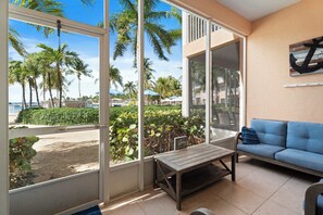 A room with a large window overlooking a beach, a blue sofa, and a wooden table. - Kai-Tastic 15B - 2BR Kaibo Condo with Pool and Beach (Rum Point)