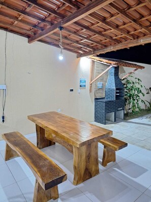 An outdoor patio with a wooden table and benches, a brick barbecue, and a ceiling with exposed beams. - Chácara Recanto do Sítio (Barbalha)