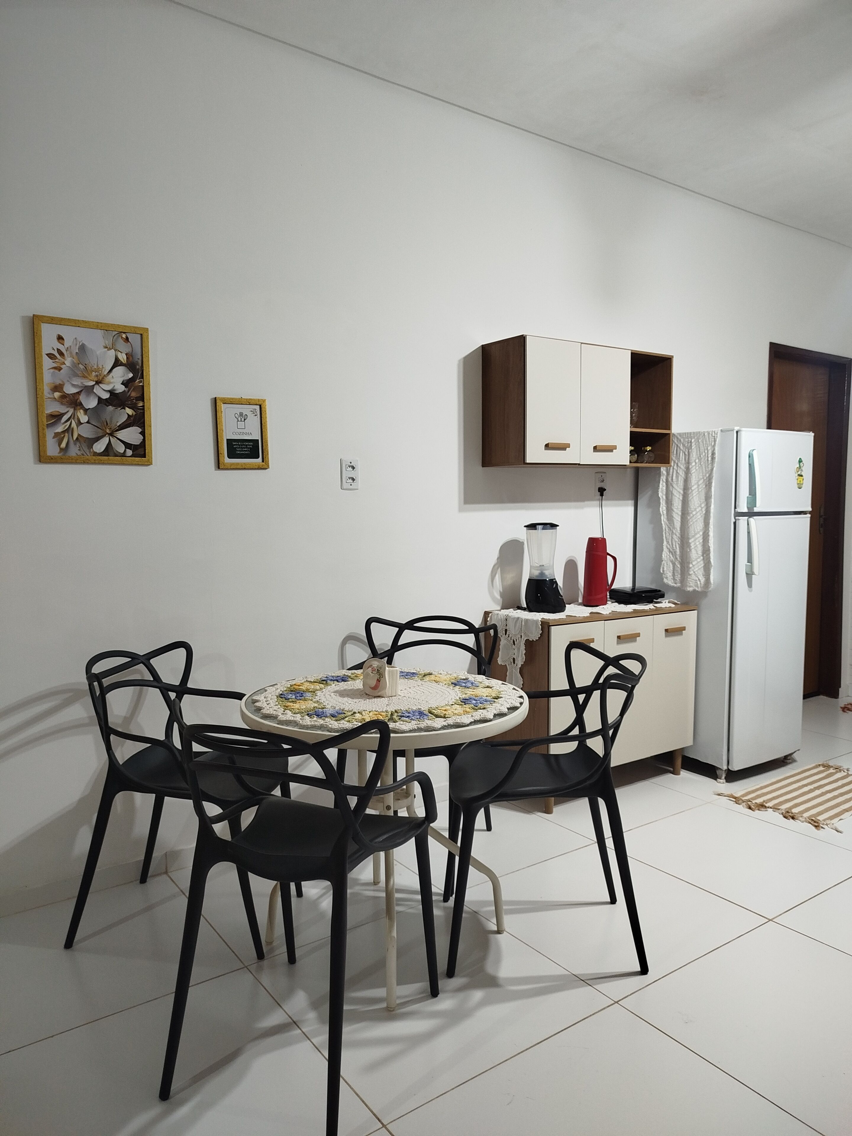 Cozinha compacta com armários brancos, uma geladeira, uma cafeteira e uma mesa com cadeiras.