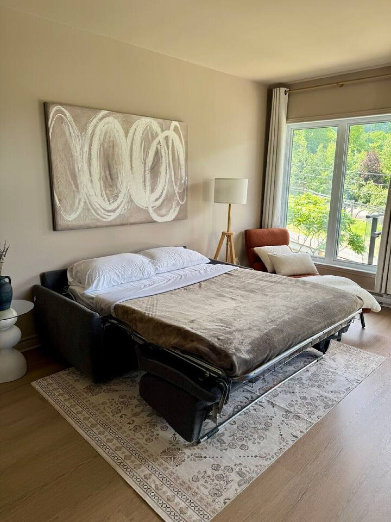 A modern bedroom with a bed, a large abstract painting, a floor lamp, and a view through a large window.