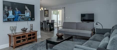 Uma sala de estar moderna com um sofá cinzento, uma TV de tela plana, um armário de madeira e uma área de jantar com mesa e cadeiras.
