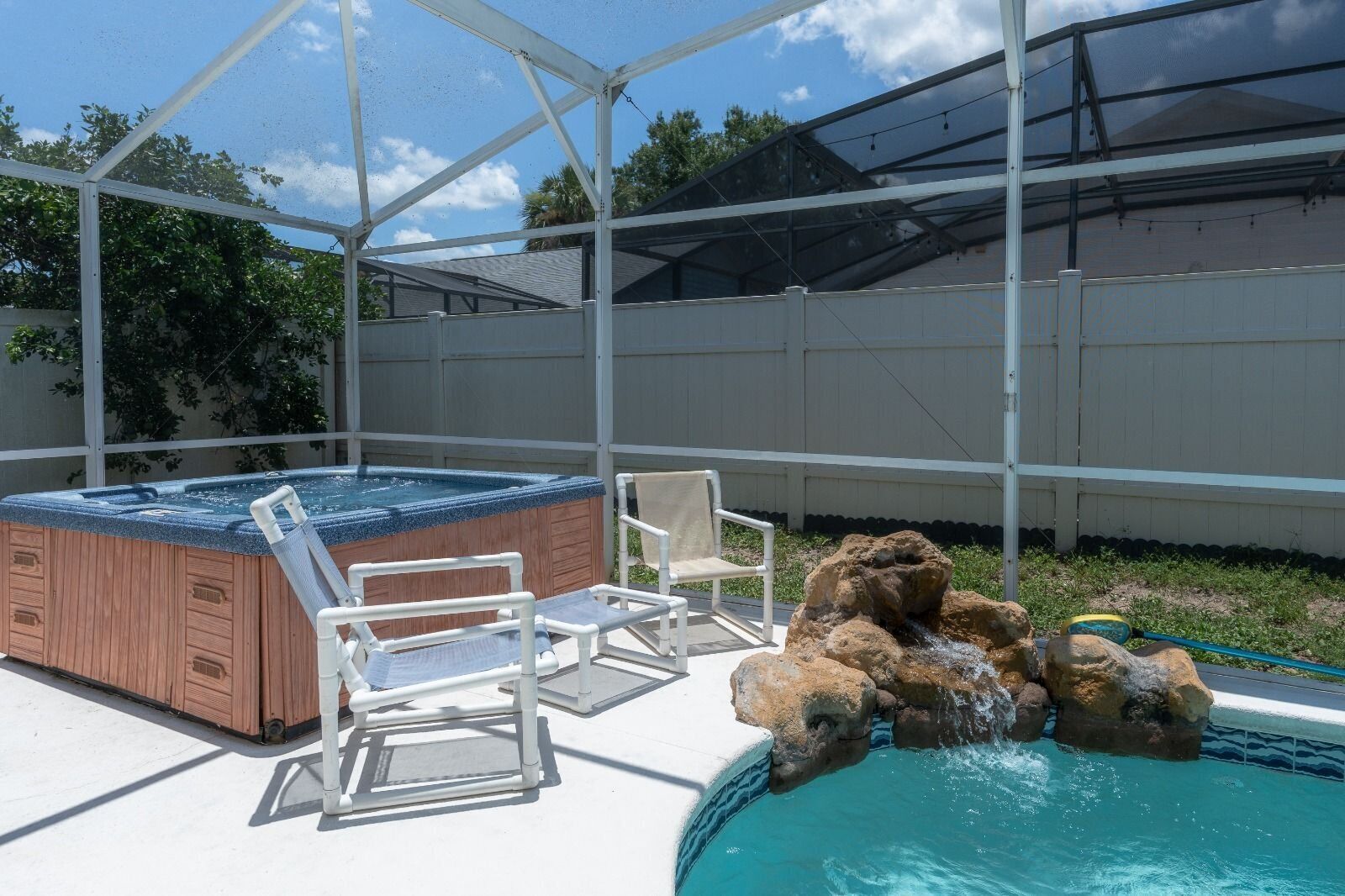 An outdoor hot tub with two chairs, a pool with a rock feature, and a covered area with a white fence.