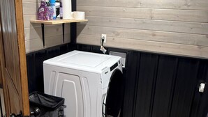 A compact laundry room with a white washer and dryer, a shelf with cleaning supplies, and a trash bin. - Lake house minutes from the boat dock w/attached garage for boats or vehicles (Foss)
