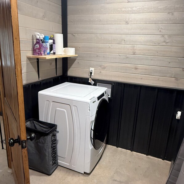 A compact laundry room with a white washer and dryer, a shelf with cleaning supplies, and a trash bin.