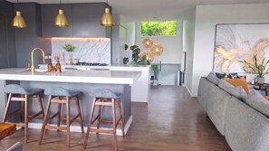 A modern kitchen with a central island, bar stools, pendant lighting, and a view of the living area with a sofa and a painting. - Luxury Hilltop Retreat: Hot Tub & Panoramic Views (Waikanae)