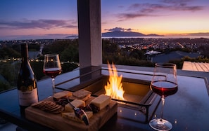 A terrace with a fire feature, a bottle of wine, glasses, and a cheese board. - Luxury Hilltop Retreat: Hot Tub & Panoramic Views (Waikanae)