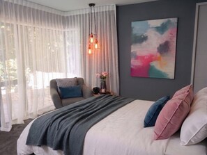 Een slaapkamer met een bed, een stoel, een nachtkastje met een vaas, en een schilderij aan de muur.