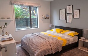 A bedroom with a bed, a desk, a window with a view of greenery, and framed artwork on the wall. - Luxury Hilltop Retreat: Hot Tub & Panoramic Views (Waikanae)
