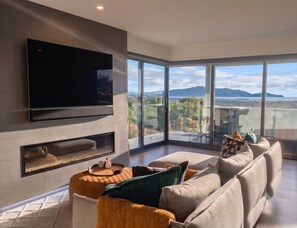 A modern living room with a flat-screen TV, a fireplace, and a large window offering a view of a lake and mountains. - Luxury Hilltop Retreat: Hot Tub & Panoramic Views (Waikanae)