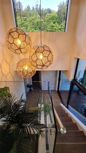A modern interior with a glass staircase, geometric pendant lights, and a view of greenery outside. - Luxury Hilltop Retreat: Hot Tub & Panoramic Views (Waikanae)