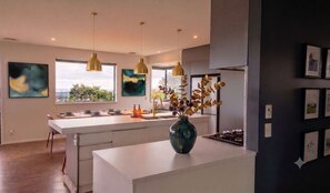 A modern kitchen with pendant lighting, a central island, and artwork on the walls. - Luxury Hilltop Retreat: Hot Tub & Panoramic Views (Waikanae)