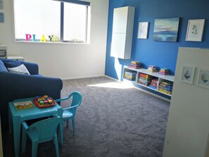 A room with a blue wall, a window, a bookshelf with board games, a blue sofa, and a small table with toys. - Luxury Hilltop Retreat: Hot Tub & Panoramic Views (Waikanae)
