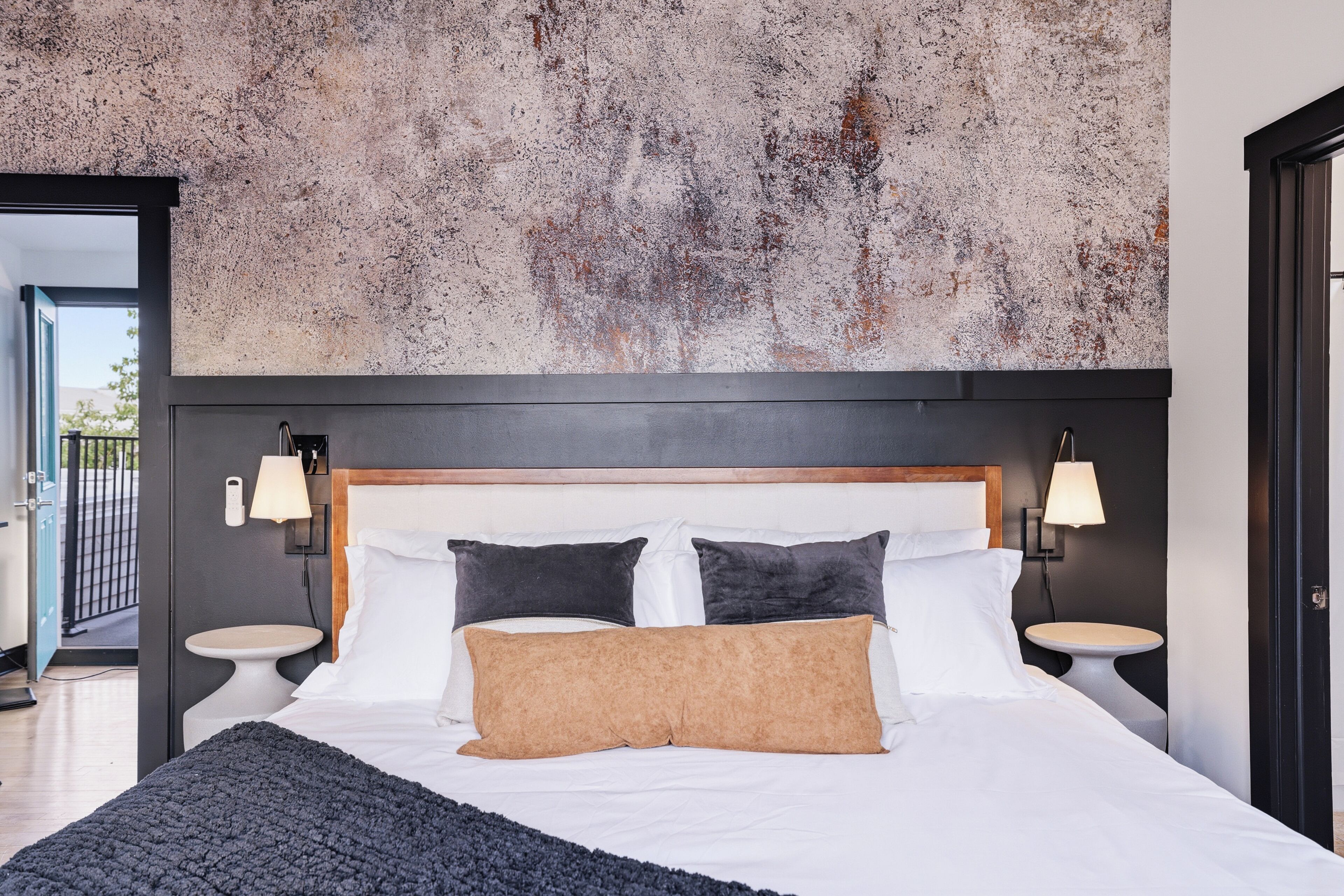 Ein modernes Hotelzimmer mit einem großen Bett, zwei Nachttischen, einer Kopfteil und einer strukturierten Wand dahinter. Blick ins Freie.