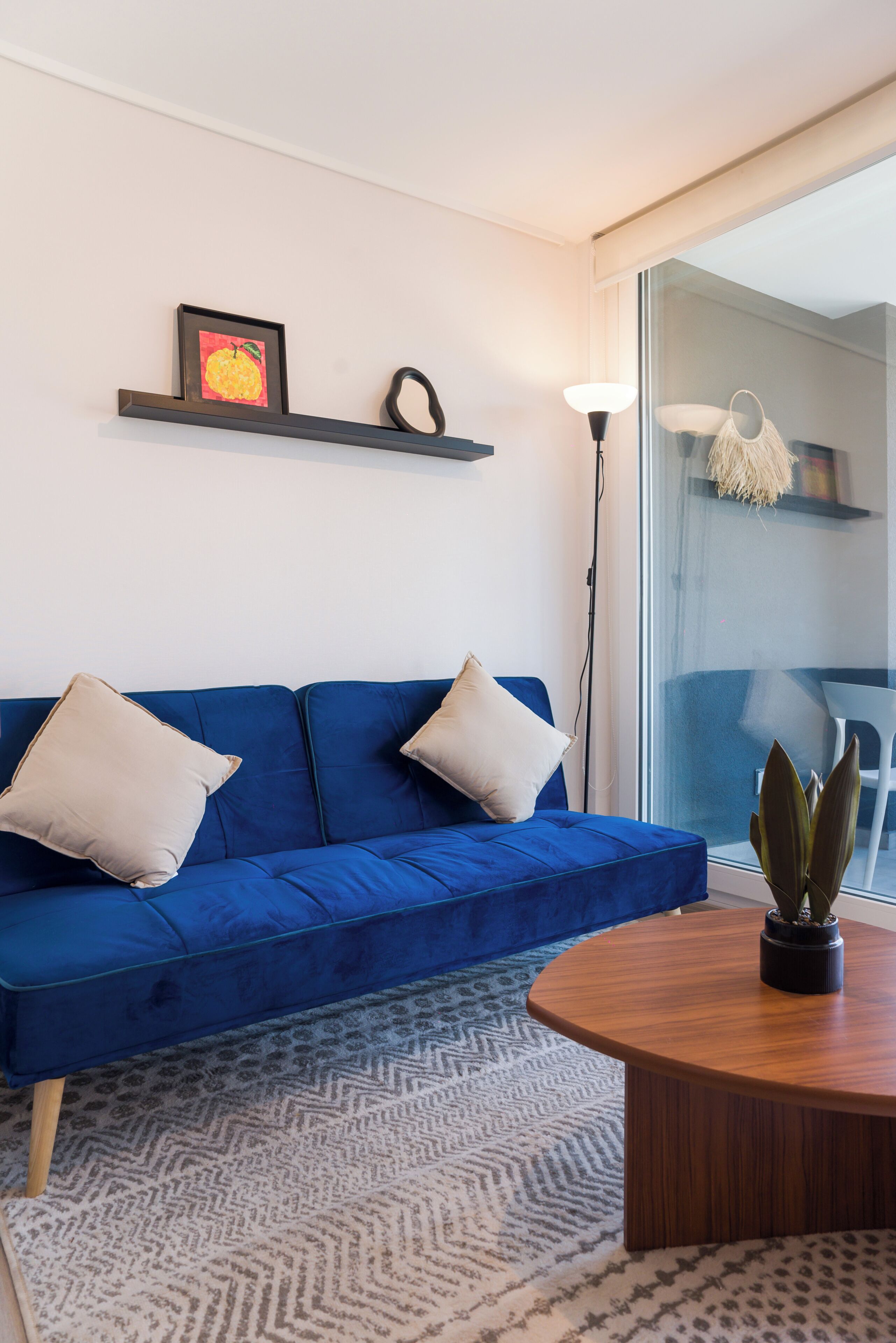 A blue sofa with two pillows, a wooden coffee table, a patterned rug, a framed picture, a floor lamp, and a potted plant.