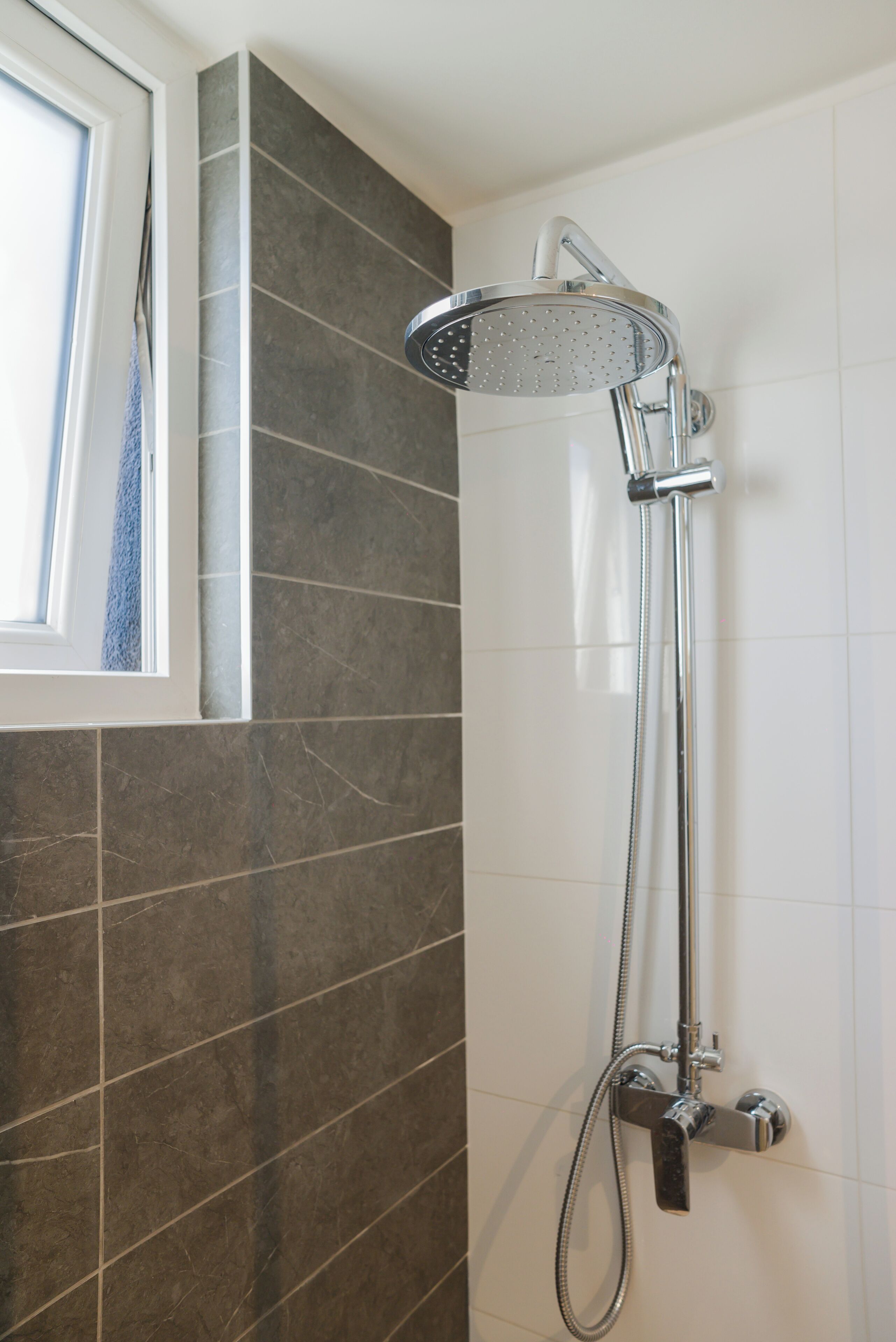 A shower with a handheld showerhead and a handheld hose, mounted on a wall with a window above it.